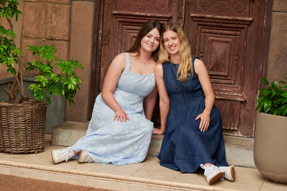 Two sisters in light blue and navy maxi dresses seated before the aged timber doors at Premier Studio