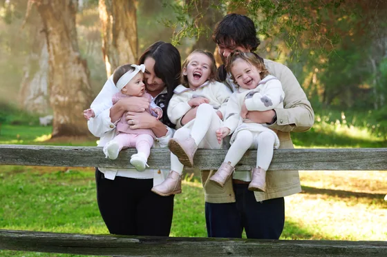 Parents in black with three little girls in cream and blush knits laughing on the timber fence at Premier Studio