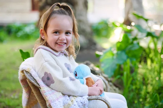 Little girl in a dusty pink knit vest and cream top cuddling a toy in the garden at Premier Studio